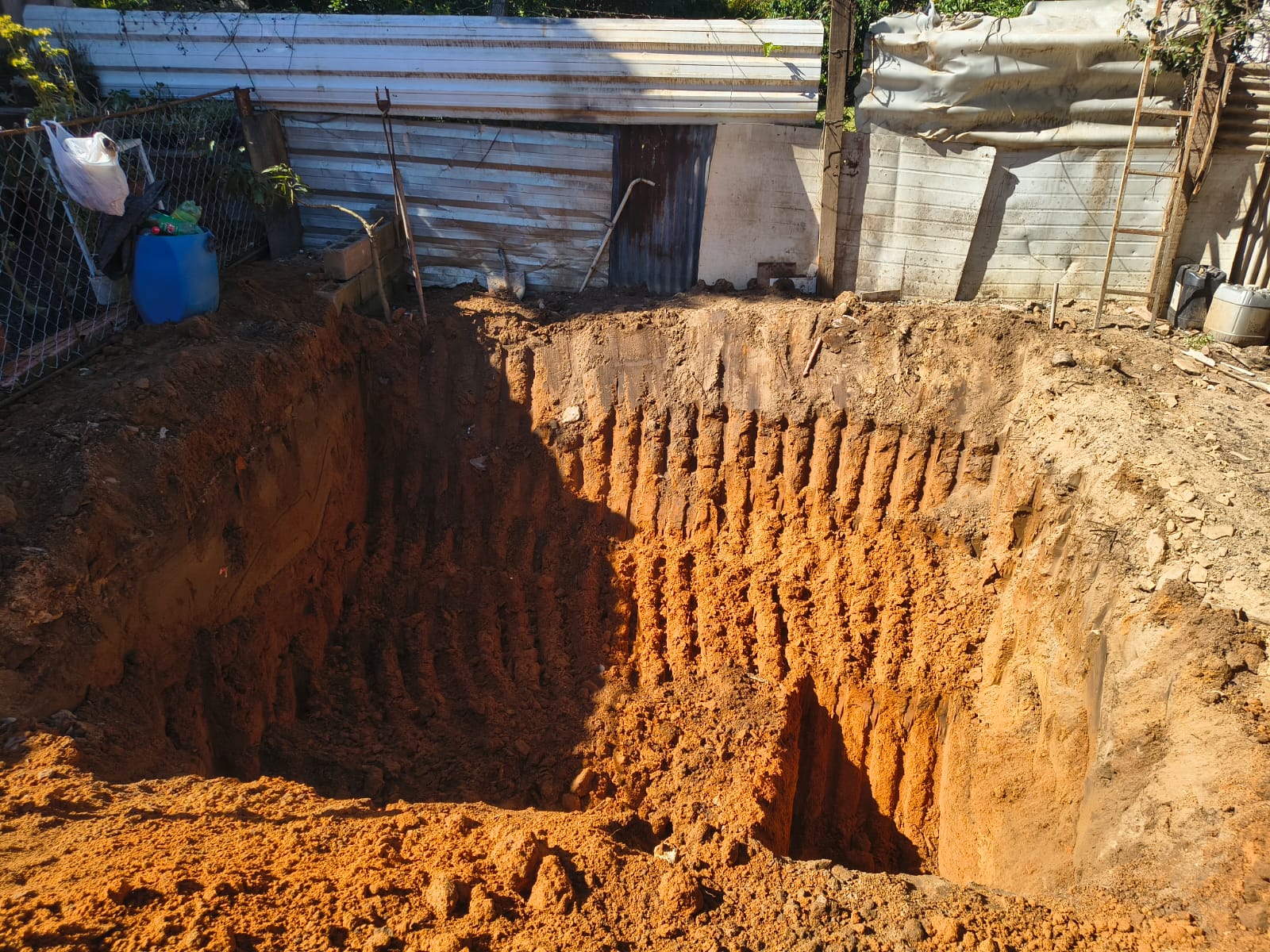 Tanque escavado em quintal murado, com as paredes de terra vermelha marcadas pela caçamba da retroescavadeira.